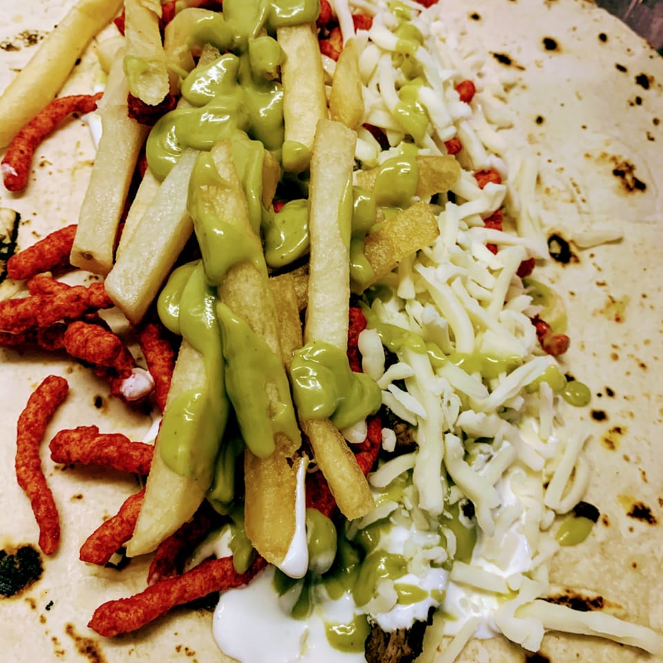 Close-up of a Wall St. Cocina burrito with fries, green sauce, shredded cheese, and red crunchy snacks.