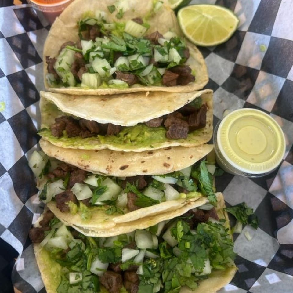 Four beef tacos with guacamole, onions, and cilantro, served with lime and sauce on checkered paper.