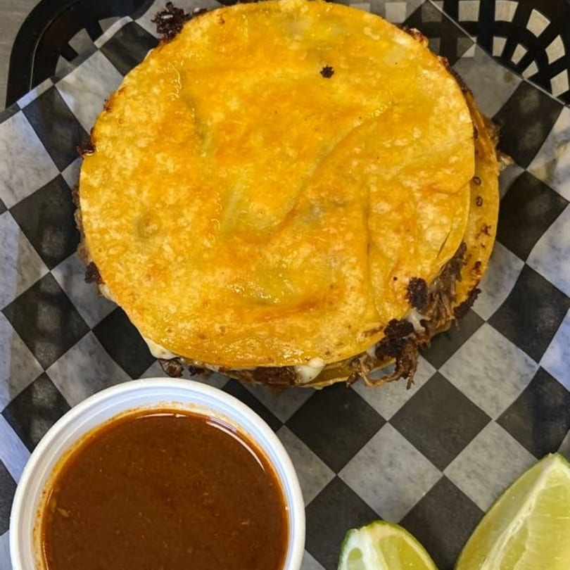 Wall St. Cocina's birria tacos with a side of consommé and lime wedges on checkered paper.